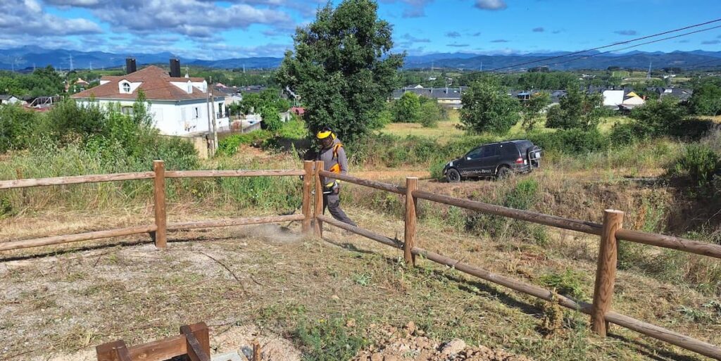 Operario realiza labores de desbroce junto a una valla de madera en San Andrés de Montejos con el paisaje del Bierzo al fondo.
