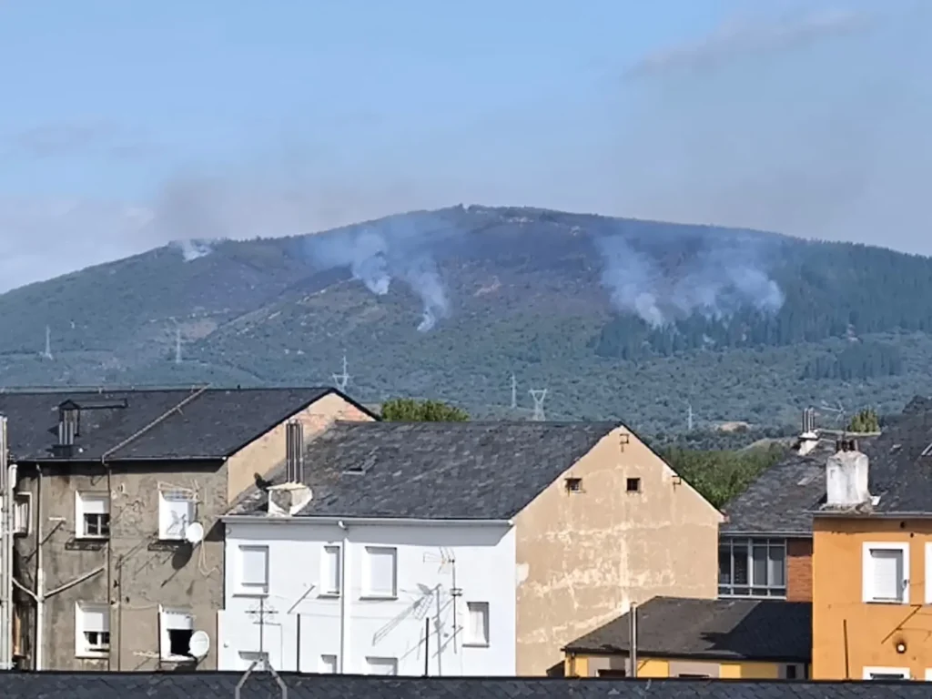 Columnas de humo en una ladera forestal afectada por los incendios, con viviendas en primer plano.